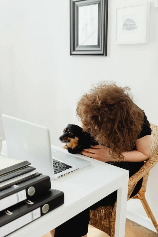 a woman sitting at a desk with her dog, while looking at a laptop