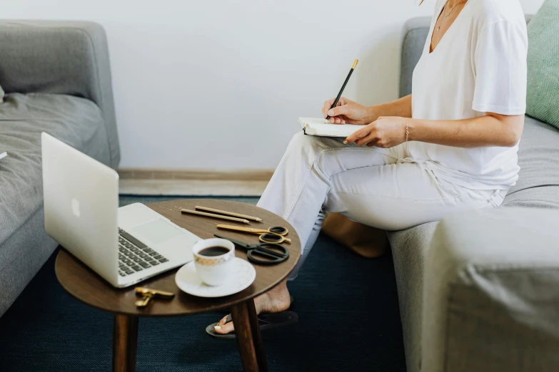 woman sitting on couch with white desk writing and laptop on table