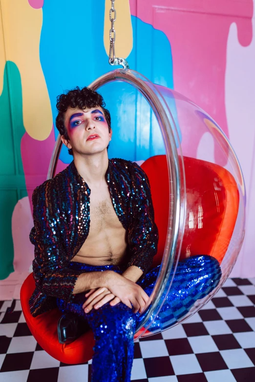 a male model wearing shiny blue pants and a shiny top sitting in a circular chair