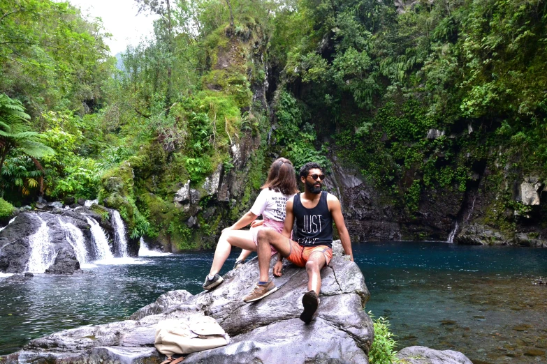 two people sitting on a rock near a small waterfall