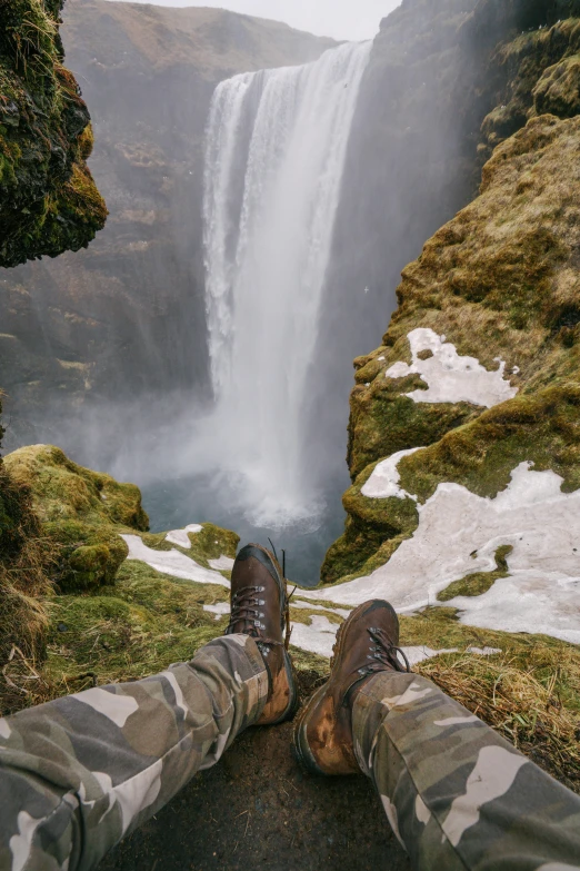 two people stand at the base of a waterfall overlooking an area with snow and snow - covered grass