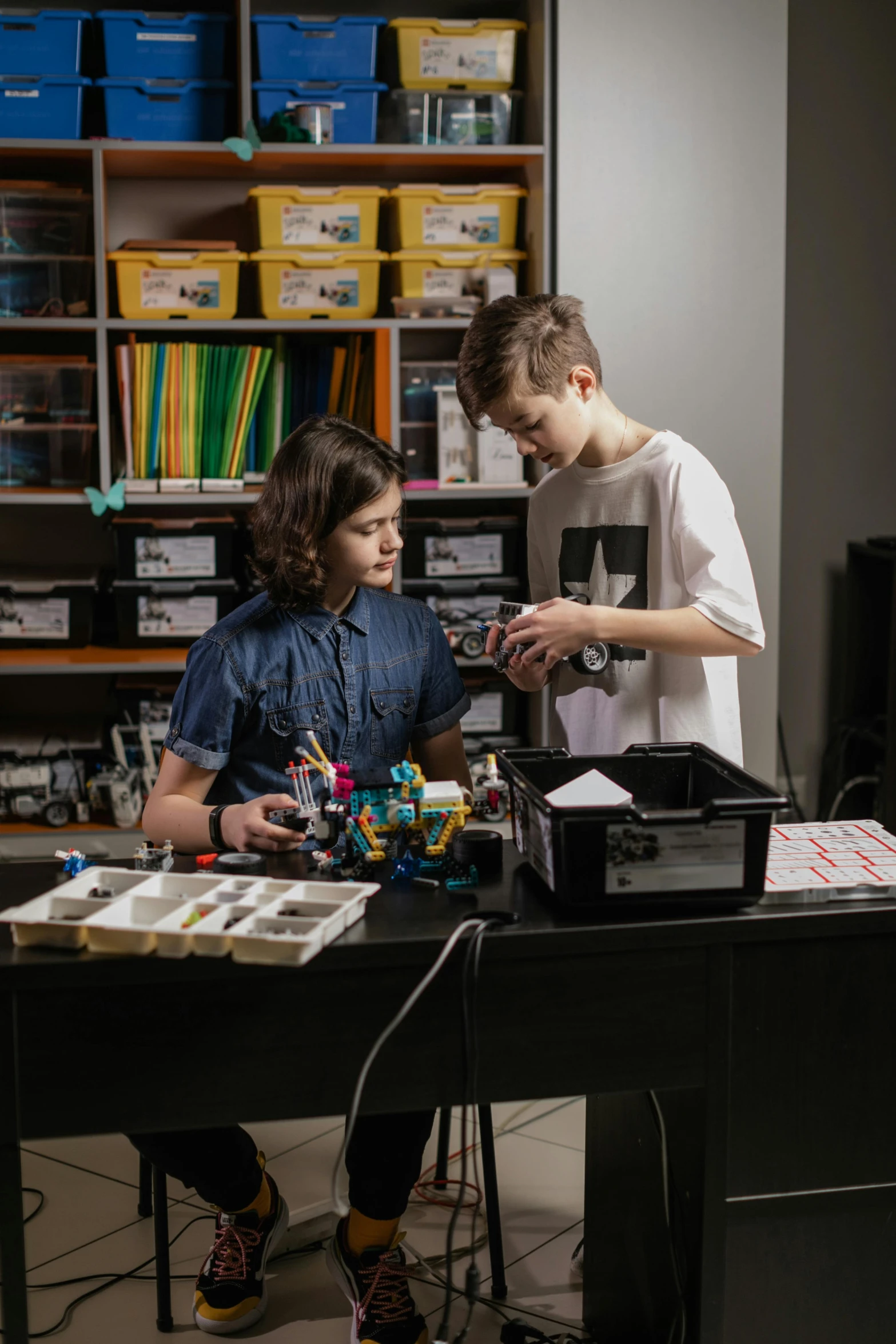 two s sitting at a black desk while a woman uses a microscope