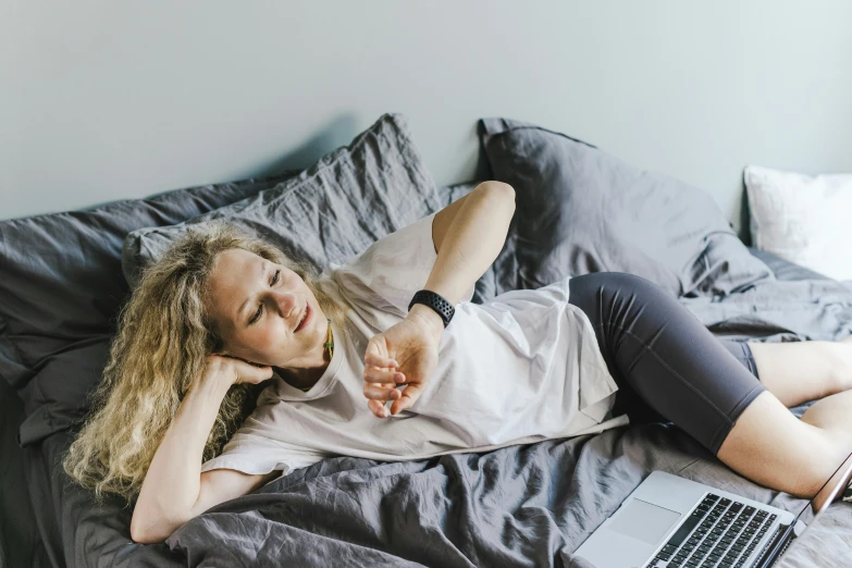 a woman in grey top laying on a bed with laptop