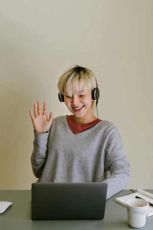 a young lady sits at a table in front of a laptop and shows off her earphones