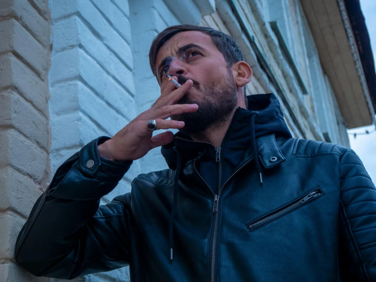 a man in a leather jacket smoking in front of a brick building