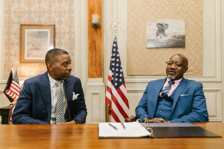 two men sit at a table in front of flags