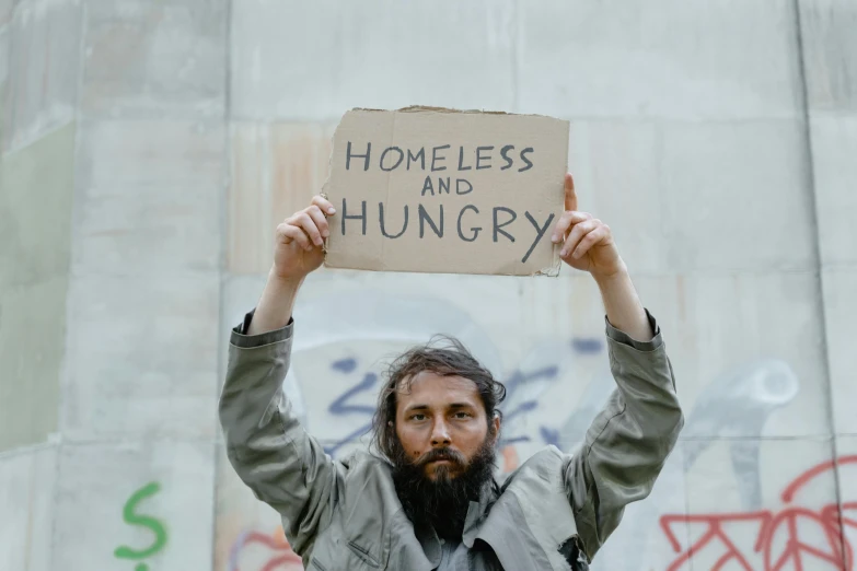 man with homeless head in front of a wall holding a cardboard sign that says homeless and hungry