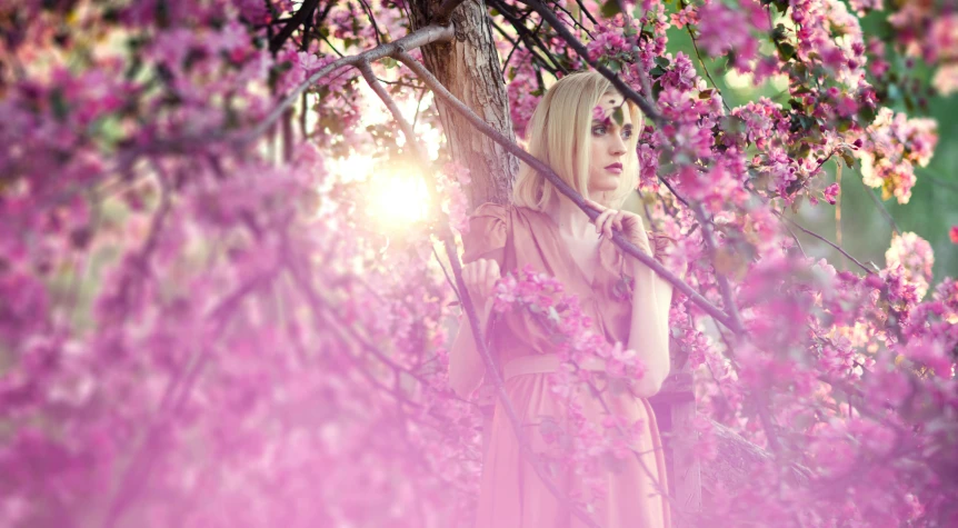 a young blonde woman wearing a pink dress stands under pink blossom trees