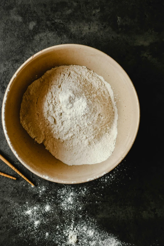 a small white bowl with flour in it on top of a dark surface