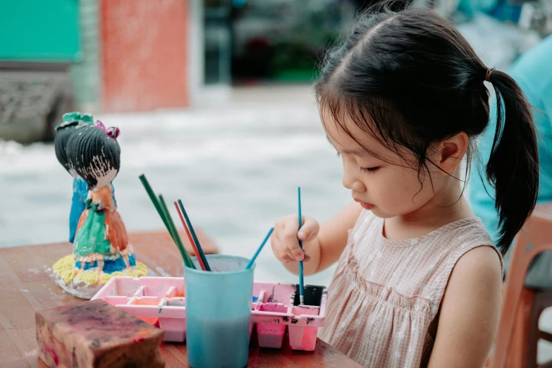 a little girl sitting at a wooden table with two brushes in her hand