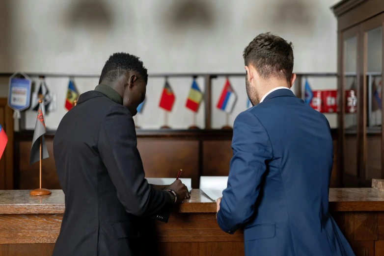 two men in suits sitting at a desk in front of flags