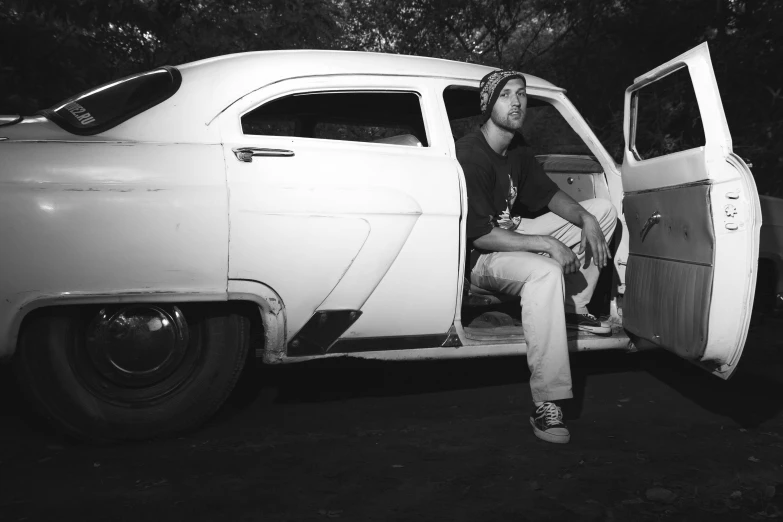 a man in cap sits on the door step of his classic car