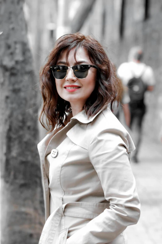 a woman with sunglasses on and a trench coat over her shoulders, is standing next to a tree in the street