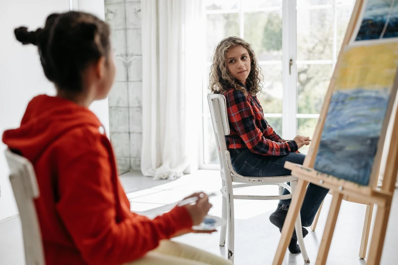 a girl is sitting in a chair while another girl is painting a picture