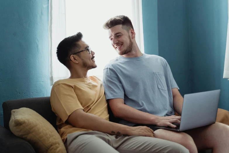 two men are on a couch one of them is sitting on a laptop