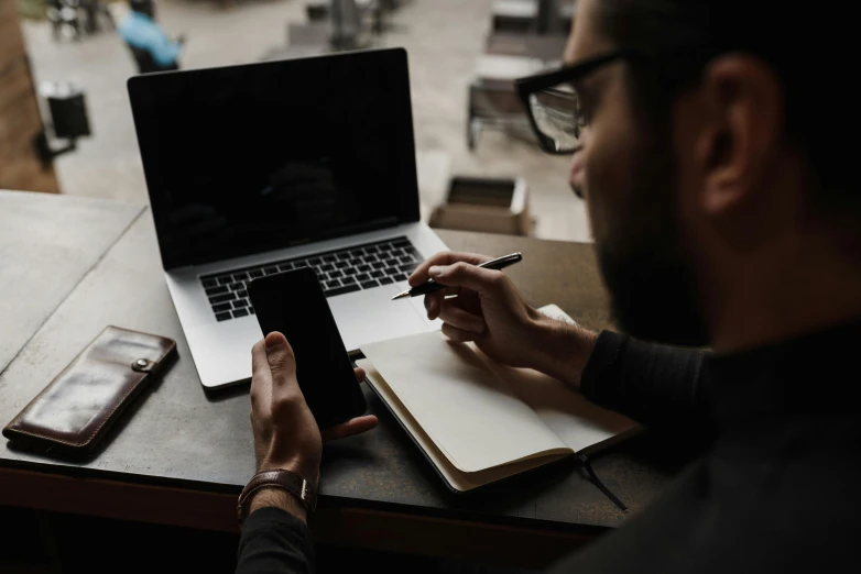 a man sitting at his laptop holding a pen and notebook in front of his