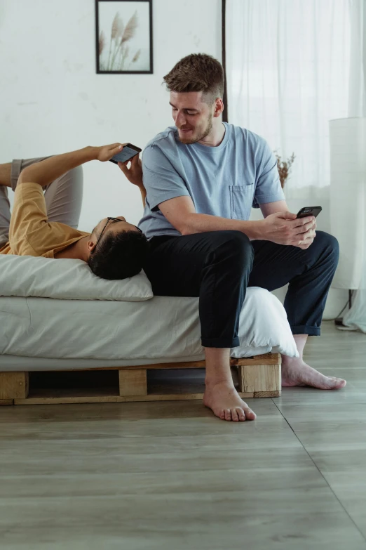 two people sitting on a bed while one looks at his cell phone