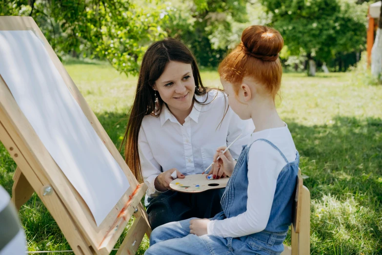 an adult sits beside a child in front of a easel holding paint