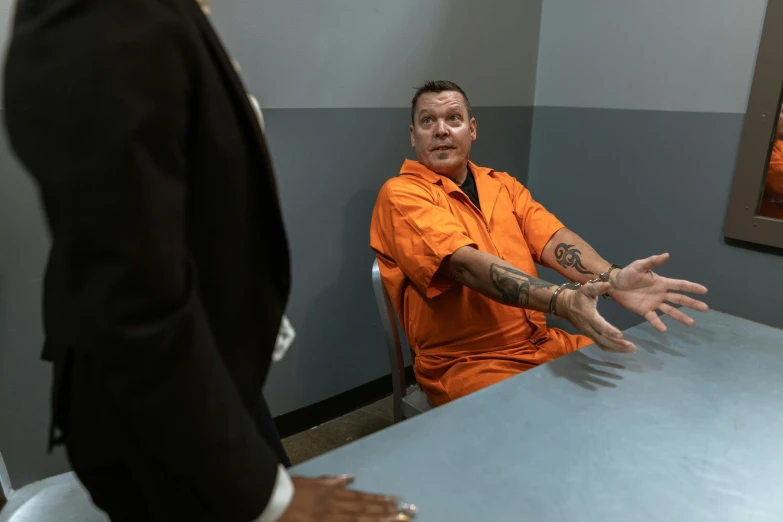 man in orange shirt and black jacket standing next to another man with his hands out