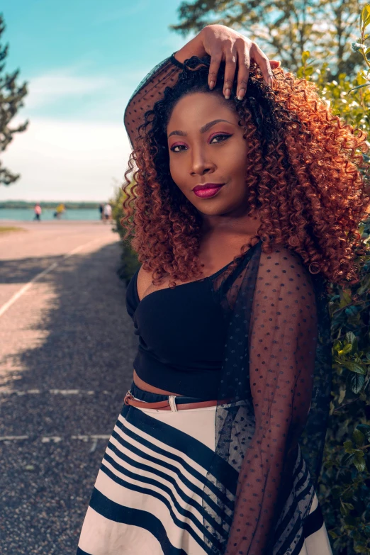 a woman wearing a black crop top and a black and white striped skirt poses by a bush