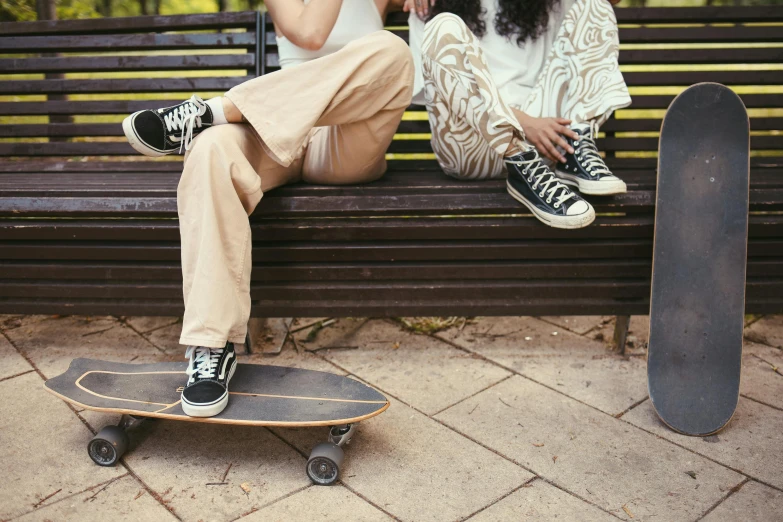 two people sit on a wooden bench, one is on a skateboard