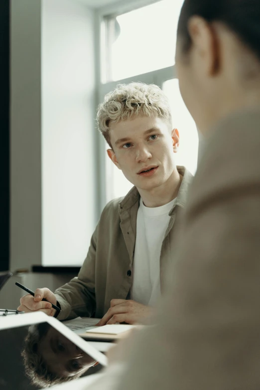 a young man sits in front of a laptop while being interviewed by a woman