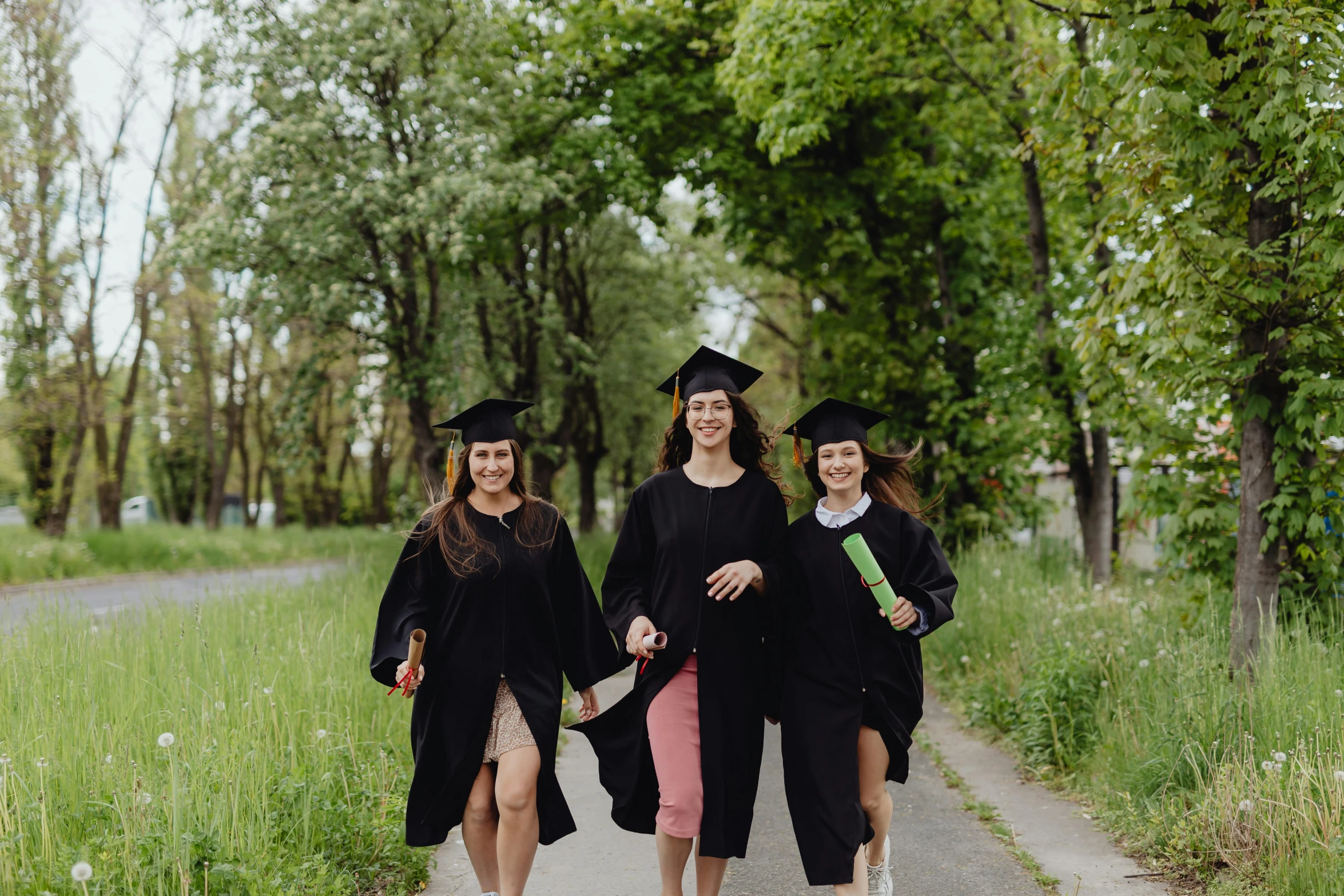 three women dressed in graduation attire walk down a path