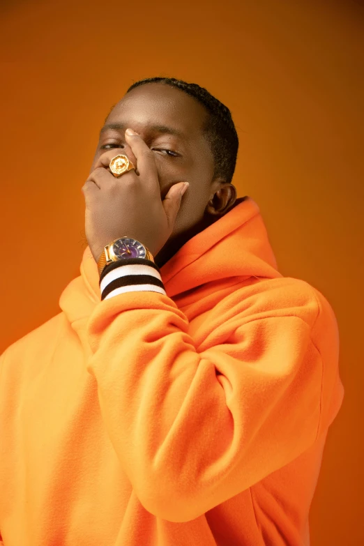 a young man standing in front of an orange wall, with his hands near his face