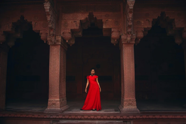 a woman in a long red dress stands in a room of bricks