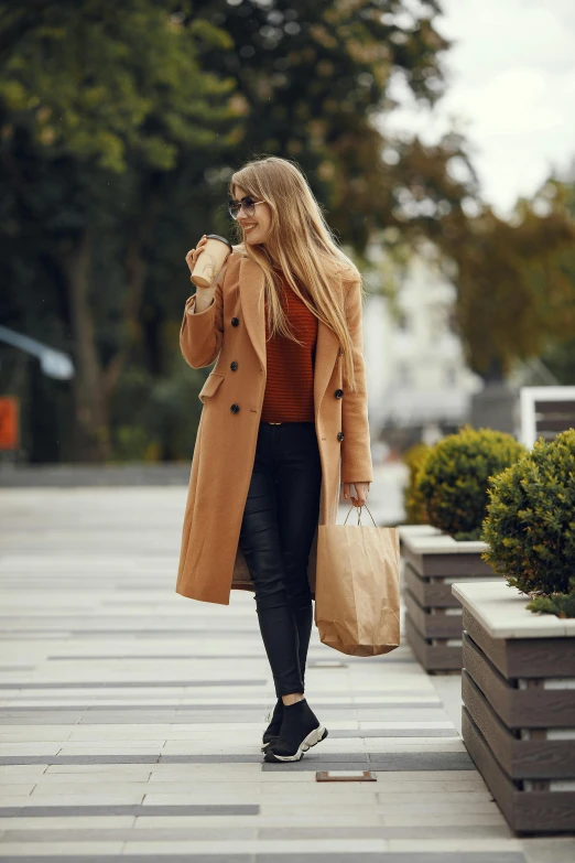 a woman in a trench coat holding two bags walks down the sidewalk