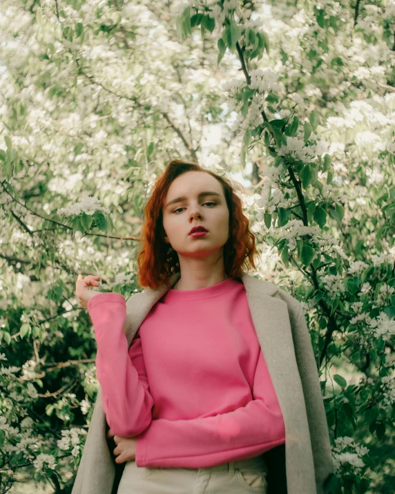 a red haired woman in a pink sweater stands next to a tree