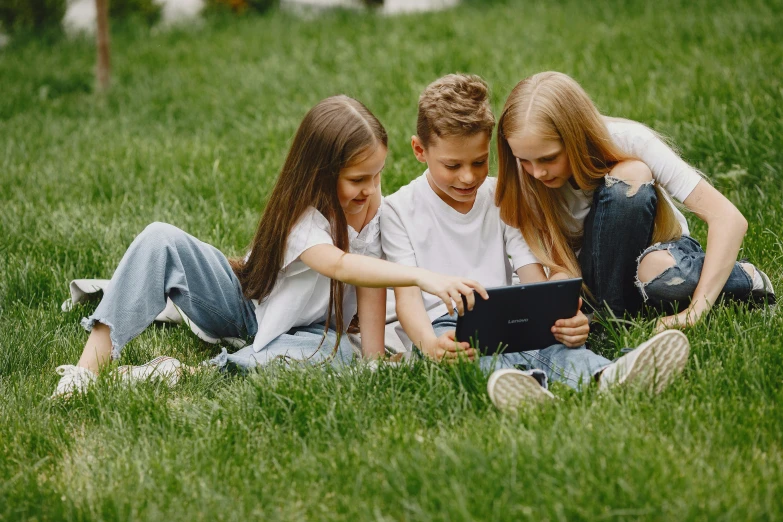 two children and one girl are sitting in a field using an electronic device