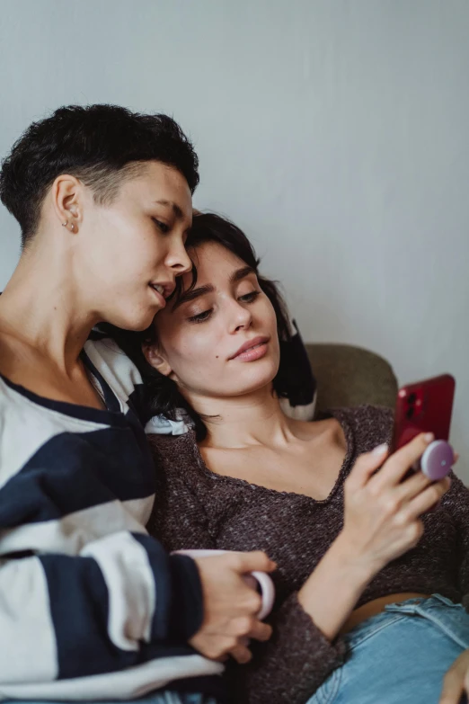 two people sitting side by side on a couch, one using a cell phone and the other holding a camera