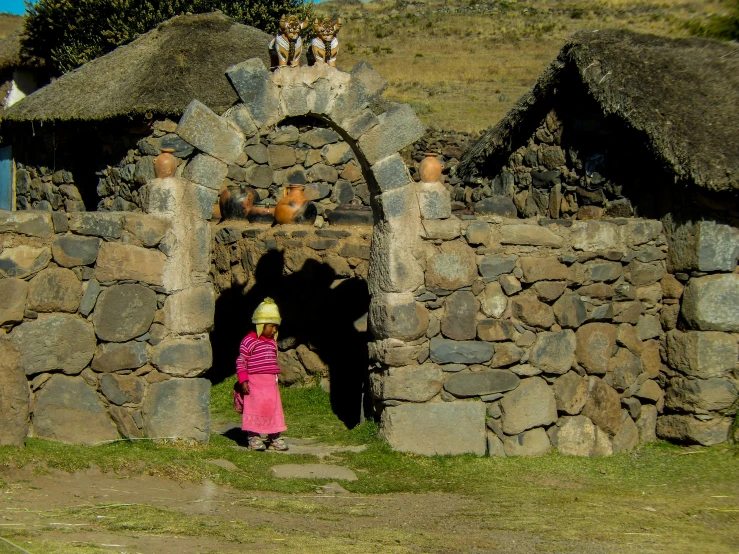 a little girl wearing a pink dress and hat in front of a stone hut