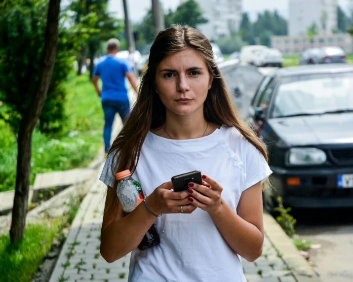 a girl in a white shirt stands on the side walk holding her phone