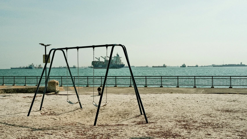 a swing set sitting on the sand with ships in the background