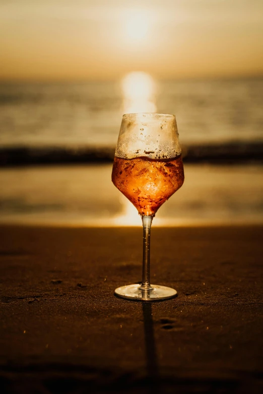 there is a wine glass with liquid sitting on the beach