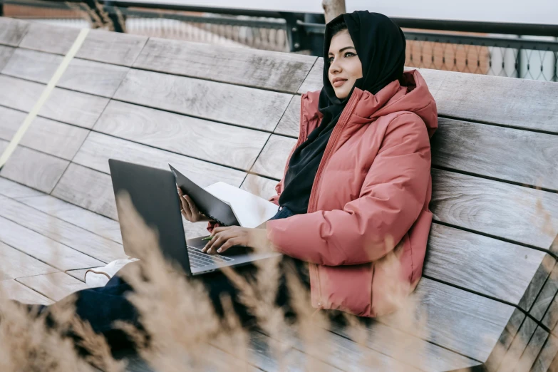 a girl is sitting on the bench and using a laptop