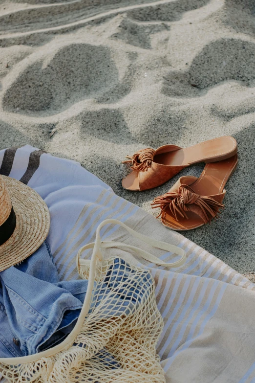 straw hat on an orange slipper, two pairs of sandals and a beach towel on a white sand background