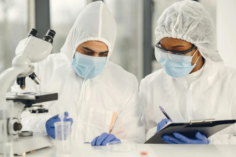 two people wearing masks, coats and lab coats are looking at a tablet while other people in protective clothing look on