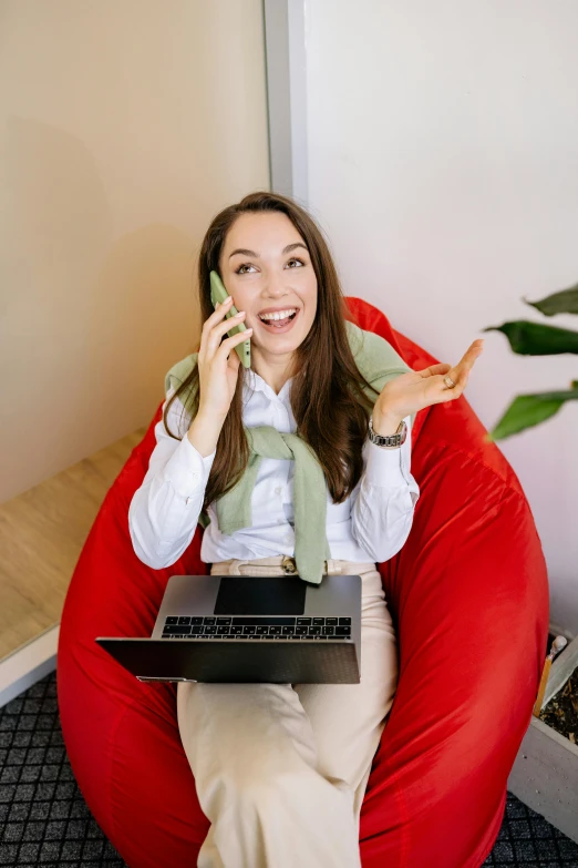 a woman sitting in a bean bag chair using a laptop while holding a phone