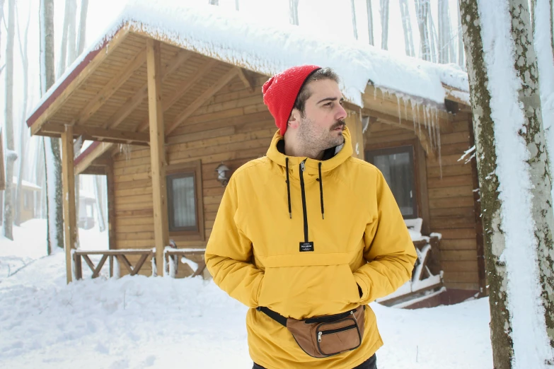 a man in a yellow hoodie and red hat standing in the snow outside a cabin