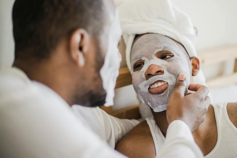 a man in a white tank top and a white towel around his head is shaving a mans face while looking into a mirror