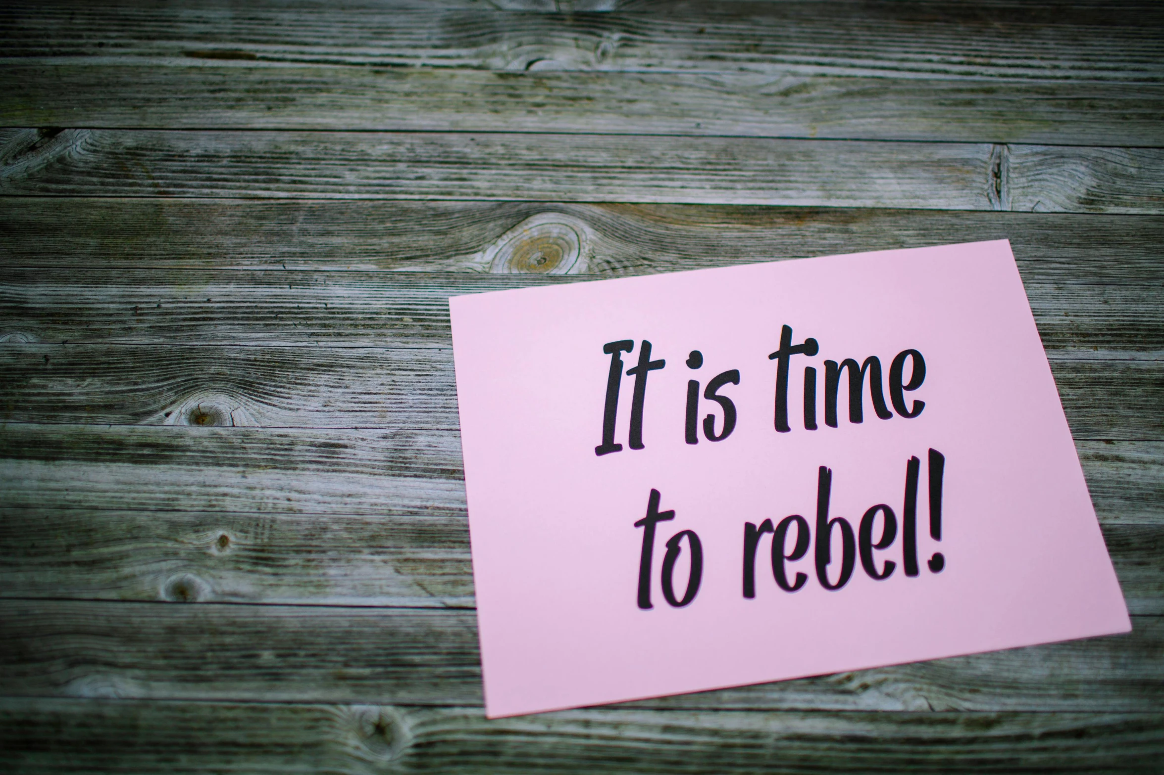 pink post it note on wooden surface it reads it is time to rebel