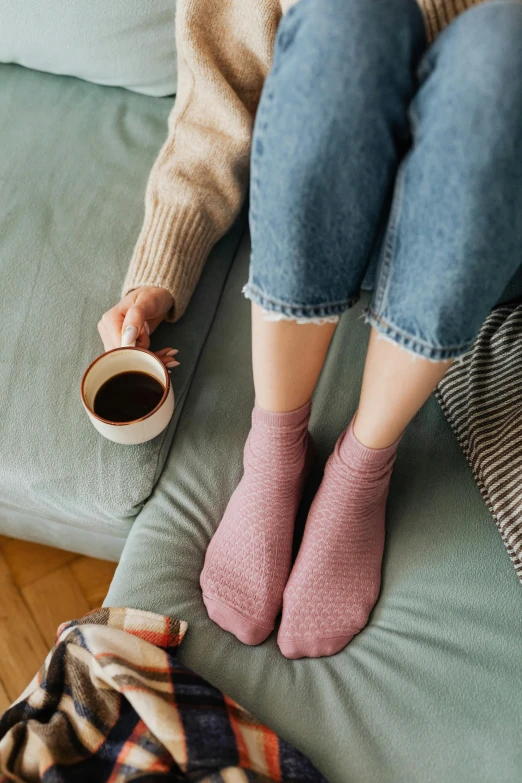 two legs with pink socks are sitting on a green couch and holding a cup of coffee