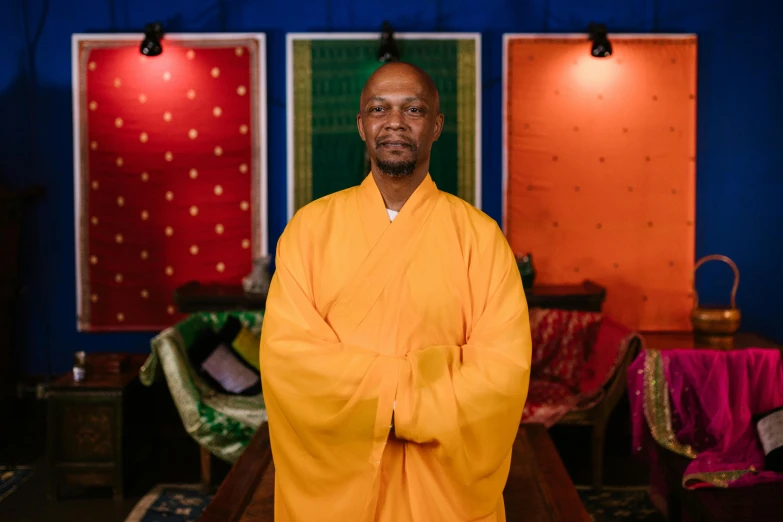 man in monk robes stands near four colorful paintings