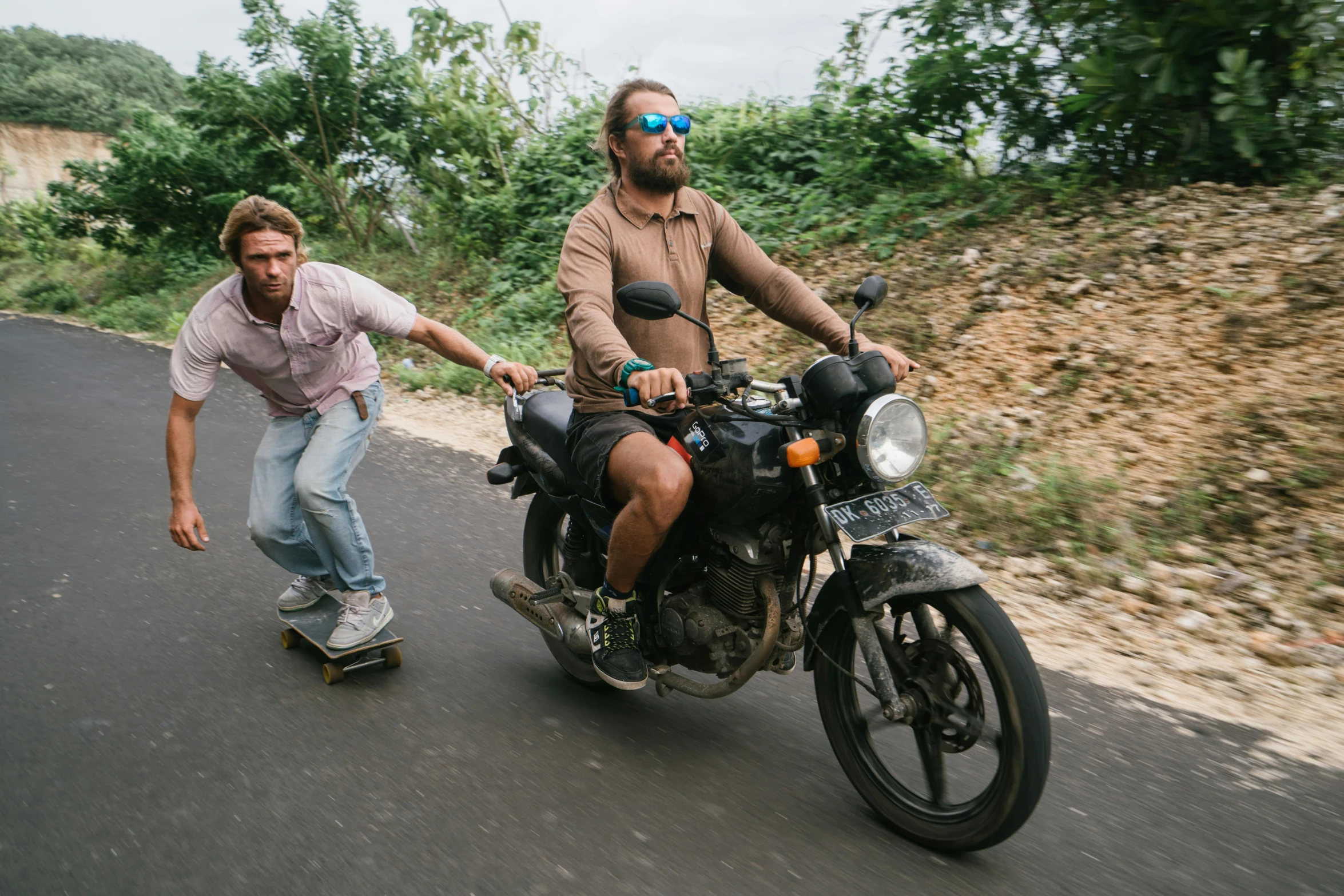 a man wearing sunglasses is riding a motorcycle with a man riding on a skateboard