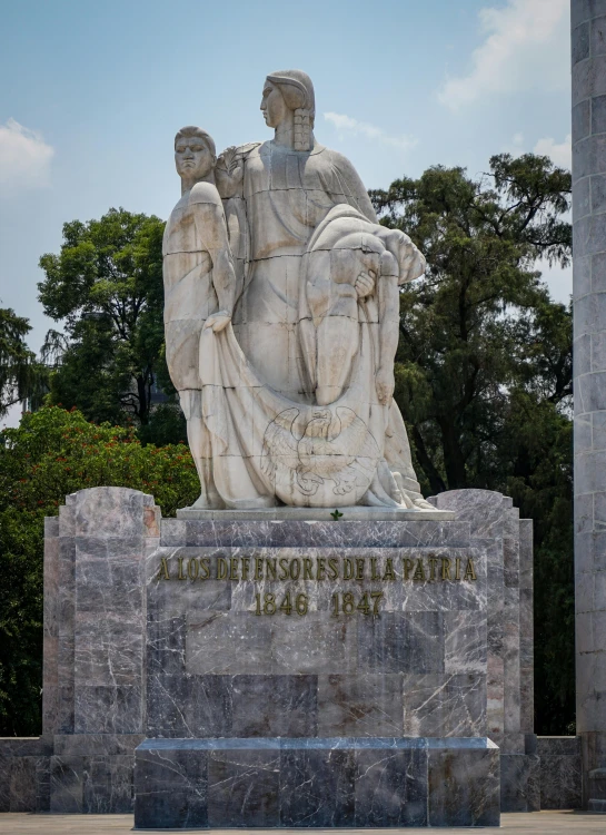 the statue shows the back of a large, seated figure with arms crossed, two other figures on either side, and trees in the background