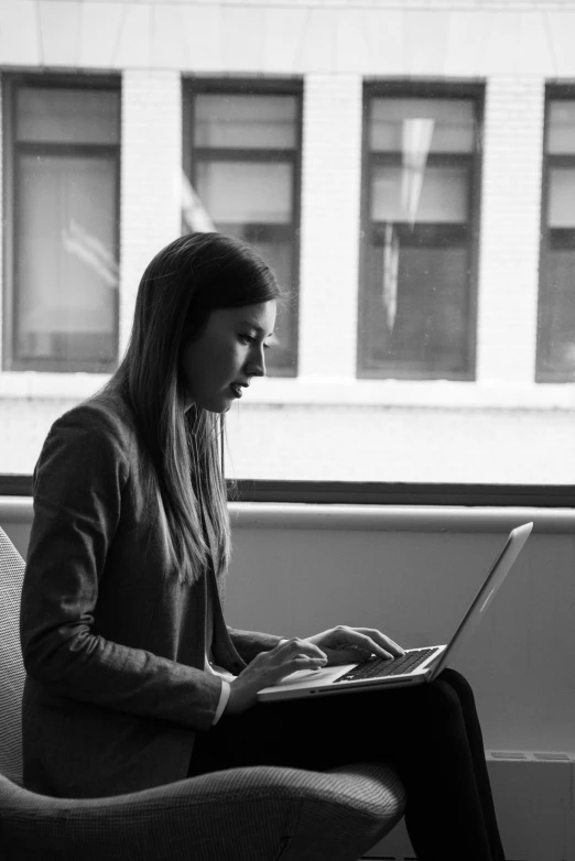 woman sitting in chair using laptop computer next to window