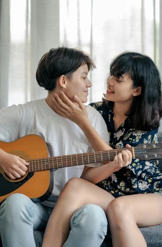 two young ladies are sitting on a couch, one woman is holding an acoustic guitar